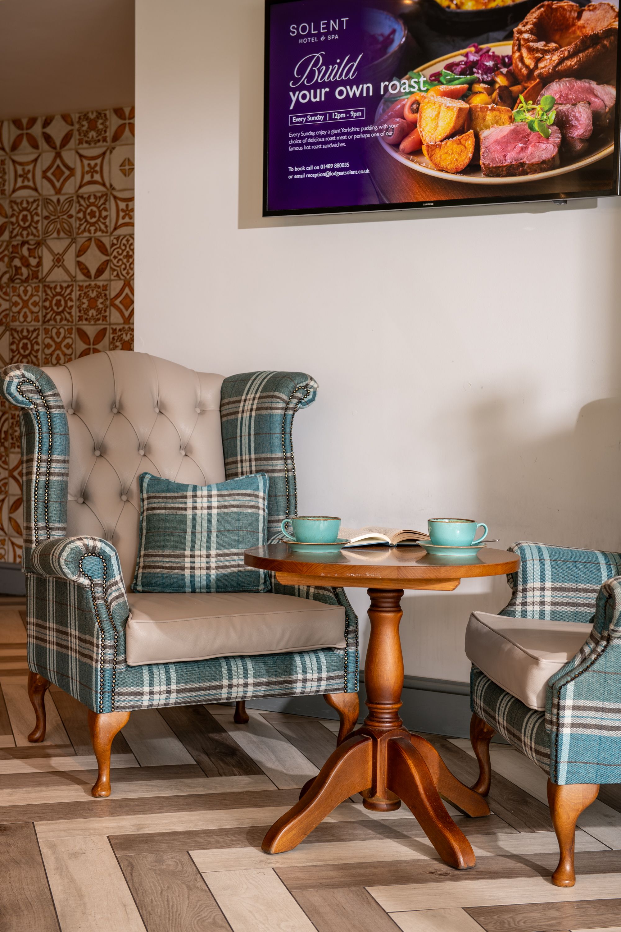 blue and cream chairs with table and coffee cups on. Behind is a TV screen showcasing hotel updates and events.